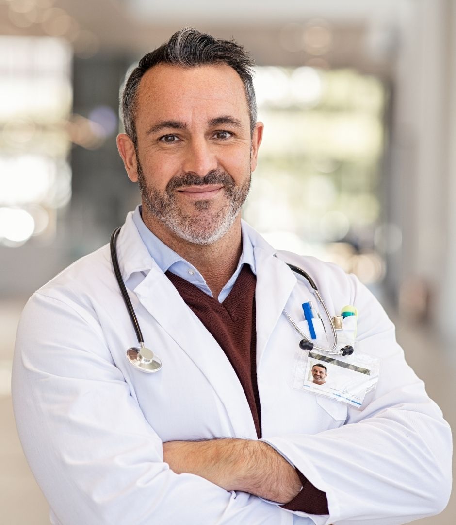 Médico de jaleco branco e estetoscópio, sorrindo de braços cruzados, representando a equipe médica que atende pela MaisMed